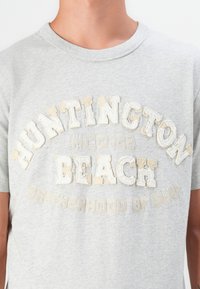 Pilkos spalvos trumpomis rankovėmis marškinėliai, papuošti tekstūriniais kreminio ir smėlio spalvos siuviniais, kurie rašo "HUNTINGTON BEACH" ir frazę "BROTHERHOOD OF SURF."