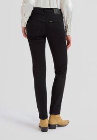 Widok z tyłu osoby noszącej czarne dopasowane jeansy, białą koszulę z długim rękawem oraz beżowe zamszowe botki na jasnym tle.