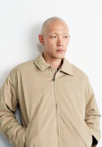 Beige lightweight jacket with a collared neckline, front zipper, and side pockets. Smooth texture and simple design with no visible patterns.