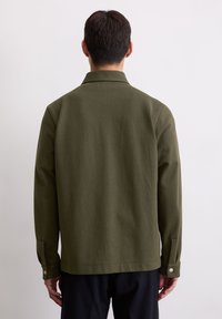 Zielona bawełniana kurtka o prostym kroju, z długimi rękawami i zapinanymi na guzik mankietami. Posiada kołnierz oraz subtelną teksturę. Pokazano widok z tyłu.