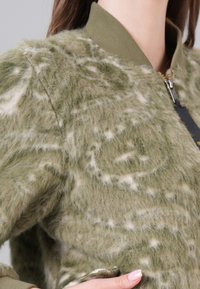 Veste bomber en matériau vert doux et duveteux avec des accents clairs, col à côtes, fermeture éclair et un motif texturé et abstrait.