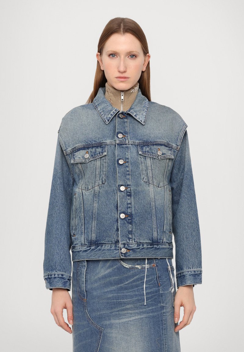 Denim jacket in light blue, featuring a collar, button closure, chest pockets, and long sleeves. Worn over a ribbed beige turtleneck.
