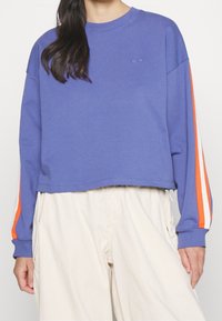 Persona che indossa un maglione corto viola con strisce arancioni e bianche sulle maniche e pantaloni beige chiaro larghi, in piedi contro uno sfondo neutro.