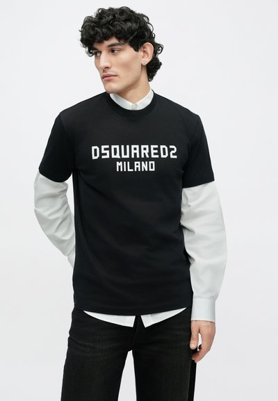 Vīrietis ar cirtainiem matiem, tērpies melnā DSQUARED2 Milano T-kreklā pār balto garroku krekla, skatās pa kreisi pret vienkāršu balto fonu.
