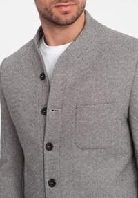 Grå herringbone-jacka med mandarin-krage, svarta knappar och en vänster bröstficka, tillverkad av texturerat tyg.