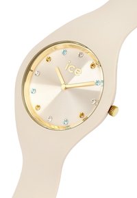 Orologio in silicone beige chiaro con cassa dorata. Quadrante argentato con indici delle ore dorati e cristalli blu, trasparenti e gialli. Corona dorata.
