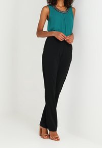 Vihreä-sininen hihaton paita, jossa on kuvioitu pääntie, yhdistetty mustiin räätälöityihin housuihin ja vaaleanruskeisiin sandaalikenkiin. Sileä kangas, kapea malli ja täyspitkä.