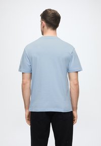 Ανοιχτό μπλε κοντομάνικο μπλουζάκι, στρογγυλή λαιμόκοψη, βαμβακερό υλικό, άνετη εφαρμογή. Φορεμένο με μαύρο παντελόνι, τα χέρια ορατά στα πλάγια.