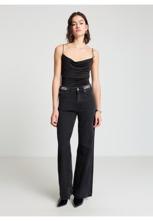 Black cowl-neck top with thin straps, paired with high-waisted, flared black jeans featuring metallic accent details on the waistband.