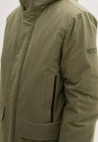 Giacca piumino verde oliva con cappuccio, dotata di chiusura con zip, due tasche laterali e dettagli neri. Tessuto liscio con una leggera trama.