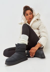 Botas de tobillo negras acolchadas con una suela gruesa y detalles de correa, combinadas con una chaqueta acolchada color crema y calcetines blancos texturizados en una figura sentada.