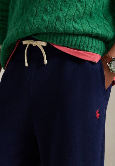 Persoon draagt marineblauwe joggingbroek met witte trekkoord, groene gebreide trui, rood overhemd eronder en polshorloge aan de linkerpols.