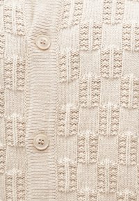 Strikket cardigan i lys beige med teksturerte firkantmønstre. Har en ribbestrikket krage og to frontknapper. Mykt, middels tykt materiale.