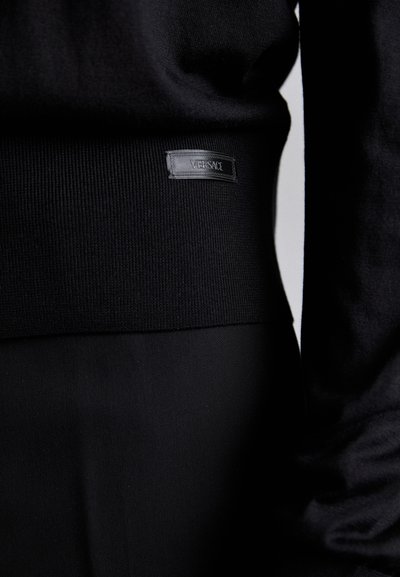 Suéter negro de punto con textura acanalada y forma ajustada. Presenta un parche con logo en el dobladillo inferior. Tela suave y diseño minimalista.