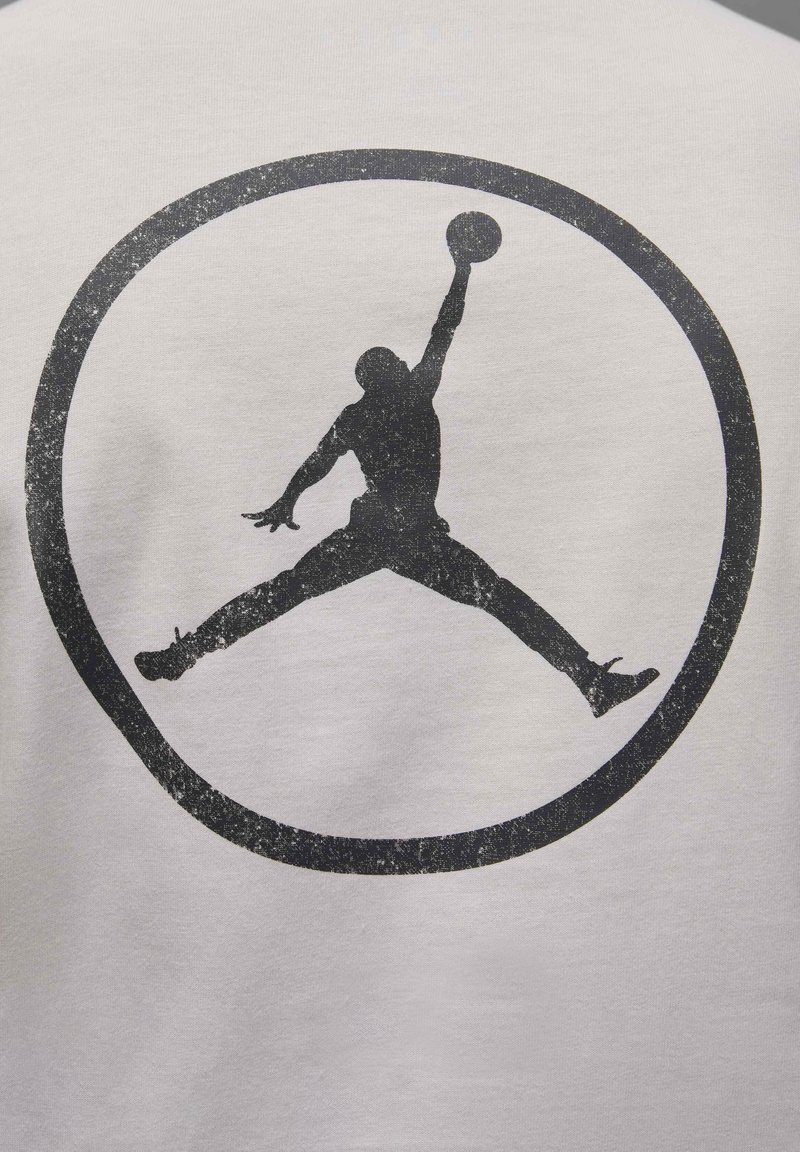 Maglietta grigia con una silhouette nera di una figura che salta con una palla da basket, racchiusa in un cerchio nero impresso.