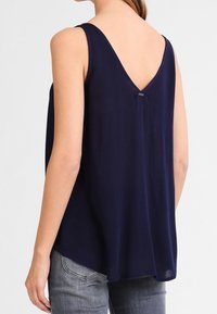 A woman wears a sleeveless navy top with a V-back, paired with light gray jeans, showcasing a simple, elegant style.