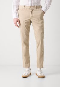 Bežīgas chinos bikses, kas izgatavotas no viegla auduma, ar rāvējslēdzēju, pogu aizdari un sānu kabatām, saskaņotas ar baltām sporta apaviem.