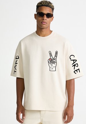 Jonge man met een zonnebril en een crème kleurig overhemd met een handgrafiek die het vredesteken toont en de woorden "Faith," "Love," "Joy," met "Take Care" op de mouwen.
