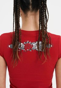 Rode, getailleerde t-shirt met een grafisch ontwerp op de achterkant, featuring schedels, een roos en florale accenten in zwart en grijs. Korte mouwen, gladde textuur.
