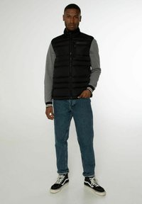 Czarna kamizelka puchowa z wysokim kołnierzem, szara koszulka z długim rękawem, niebieskie dżinsy oraz czarne trampki za kostkę. Szczegół poziomego pikowania.