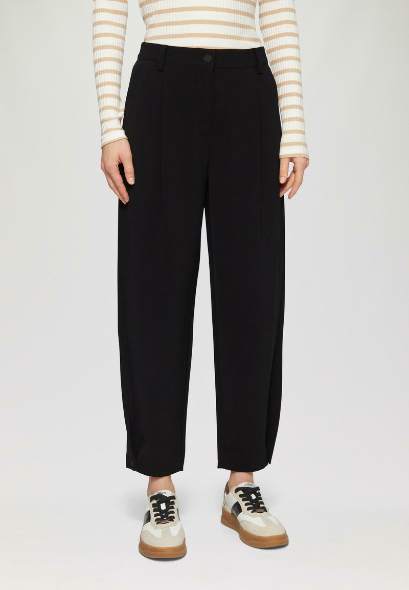 Zwarte wijde broek met voorplooien, voorzien van een knoopsluiting en zakken. Gepaard met witte en beige sneakers met zwarte accenten.