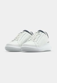 Λευκά αθλητικά παπούτσια χαμηλής κορυφής με μαύρες ταμπέλες στη φτέρνα και κορδόνια, με την επωνυμία Valentino, παρουσιάζονται σε απλό φωτεινό φόντο.