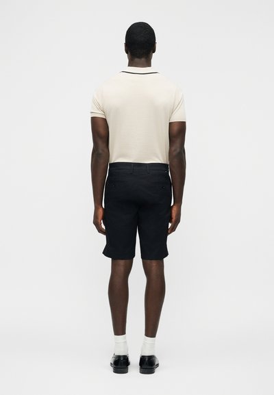 Homme portant un polo beige à manches courtes, un short noir jusqu'aux genoux, des chaussettes blanches et des chaussures noires, debout, face arrière, sur un fond uni.