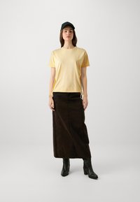 Gelbes Kurzarm-T-Shirt mit seitlichen Bindungen, kombiniert mit einem braunen Denim-Maxikleid und schwarzen Absatzstiefeln. Das Model trägt eine schwarze Mütze.