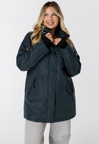 Donkerblauwe geïsoleerde jas met een hoge kraag, ritssluiting zakken en zwarte duimgatenmanchetten. Glad materiaal, gewatteerd ontwerp, knielengte.