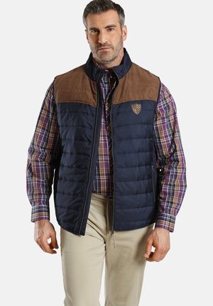Navy Puffer-Weste mit braunen Wildleder-Schultern, Reißverschluss und Logo-Patch. Über einem karierten Hemd getragen, das eine strukturierte Gestaltung und Textur aufweist.