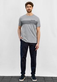 Šedé tričko s krátkým rukávem a vodorovnými černými pruhy, s kapsou. V kombinaci s námořnickými cargo kalhotami a tmavými teniskami. Model stojí na dlaždicovém povrchu.