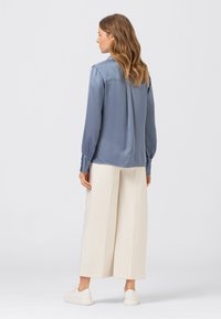 Kvinna med vågigt hår som bär en blå långärmad blus, krämfärgade vida byxor och vita sneakers, stående med ryggen mot en enfärgad bakgrund.