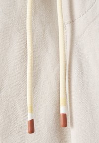 Tissu beige avec un motif chevron, équipé de deux cordons de serrage. Les cordons sont crème avec des embouts marron et de petits accents blancs.