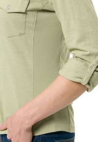 Blusa verde chiaro con un davanti liscio e pannelli laterali a coste. Presenta polsini con bottoni e un collo a giro, realizzata in tessuto morbido.