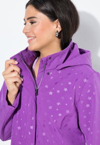 Nainen hymyilee, pukeutuneena purppuraan takkiin, jossa on tähtikuvioita ja hopeiset renkaat, kiinnittäen kauluksen vetoketjua.