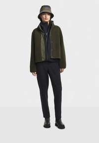 Zelena flis jakna s kontrastnim olivno zelenim sprednjim delom, kombinirana s črno majico in oprijetimi črnimi hlačami. Črne gležnjarice dopolnjujejo videz.