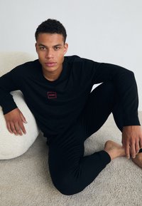 Cămașă neagră cu mâneci lungi, având un mic logo roșu dreptunghiular și pantaloni negri ajustați. Textură netedă, așezat pe o suprafață pufoasă de o culoare deschisă.