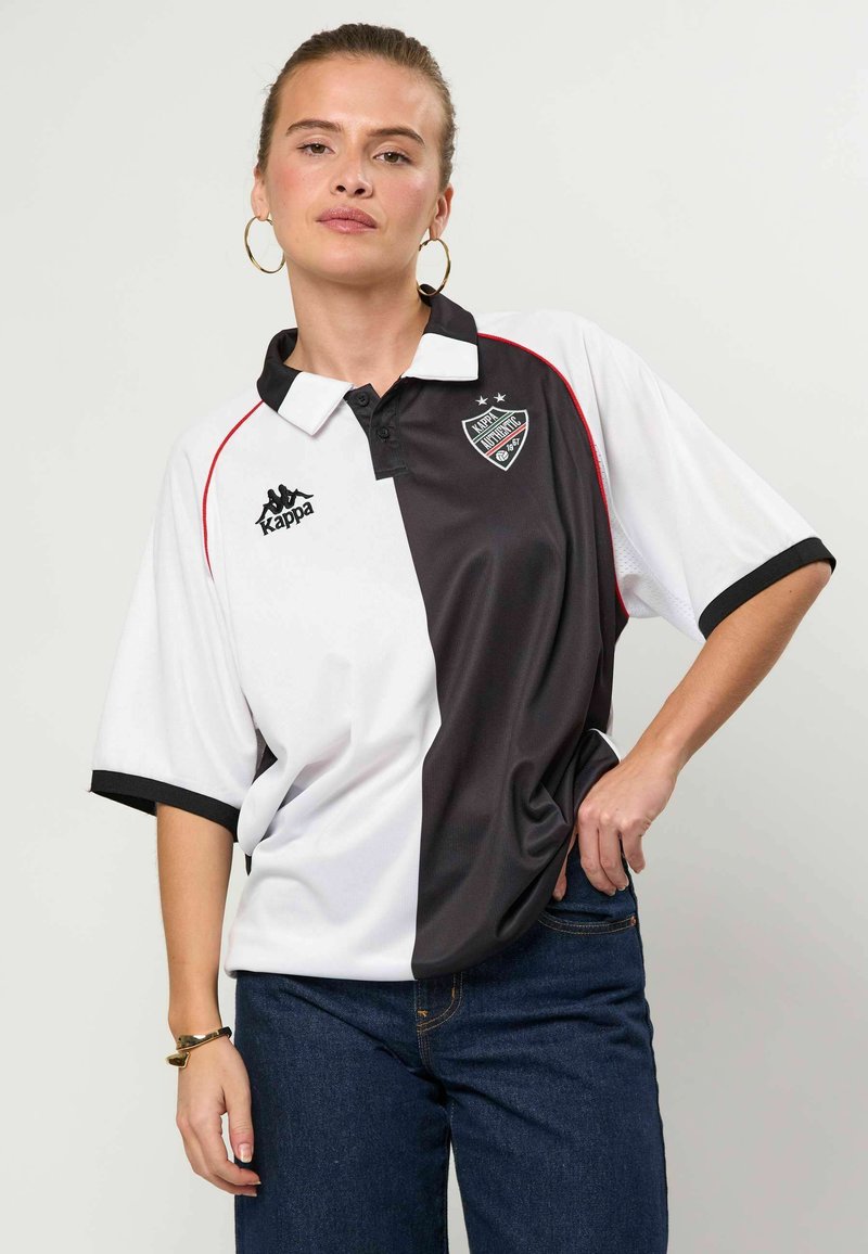 Jeune femme portant des boucles d'oreilles créoles et un polo Kappa bicolore noir et blanc avec des logos, posant avec une main sur la hanche et un jean.