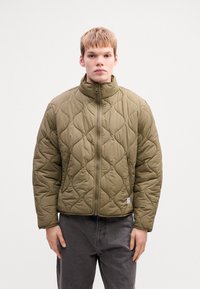Joven vestido con una chaqueta acolchada verde oliva con cremallera frontal y bolsillos con cremalleras, combinada con pantalones gris oscuro, de pie frente a un fondo blanco.