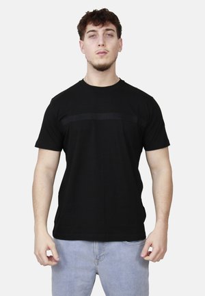 Uomo con capelli corti e ricci che indossa una t-shirt nera a maniche corte e jeans azzurri, in piedi davanti a uno sfondo bianco.