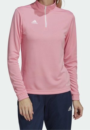 Kobieta ubrana w różową, sportową bluzę z długim rękawem i suwakiem do ćwierci wysokości oraz granatowe spodnie Adidas, stojąca z rękami swobodnie opuszczonymi wzdłuż ciała.