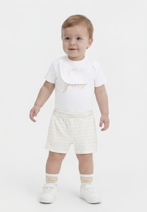 Småbarn iført hvit kortermet skjorte, matchende shorts, smekke, hvite sokker og joggesko, stående mot en ensfarget hvit bakgrunn.