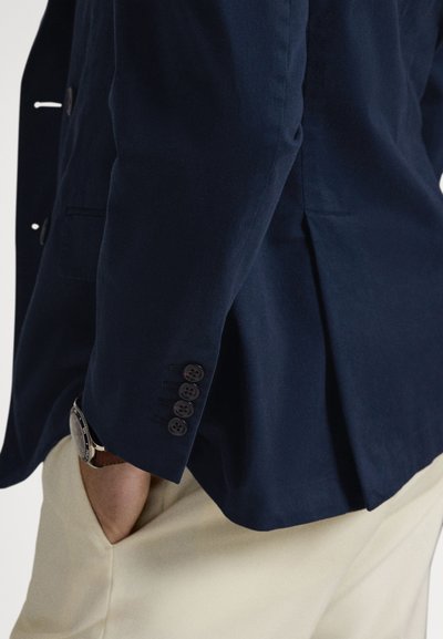 Tamsiai mėlynas švarkas su lygiu paviršiumi, prigludusiu siluetu ir penkiais mygtukais ant rankogalių. Pateiktas su kreminės spalvos kelnėmis.