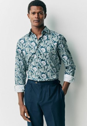 Camicia a stampa floreale in tonalità di teal e panna, con maniche lunghe e colletto button-down. Abbinata a pantaloni a pieghe blu navy.