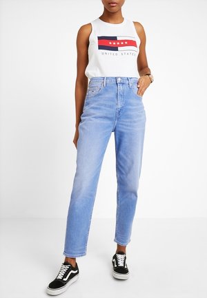 Kobieta w białej koszulce bez rękawów z czerwonym i granatowym wzorem gwiazdy oraz napisem, jasnoniebieskich jeansach z wysokim stanem i czarnych tenisówkach z białymi sznurowadłami.