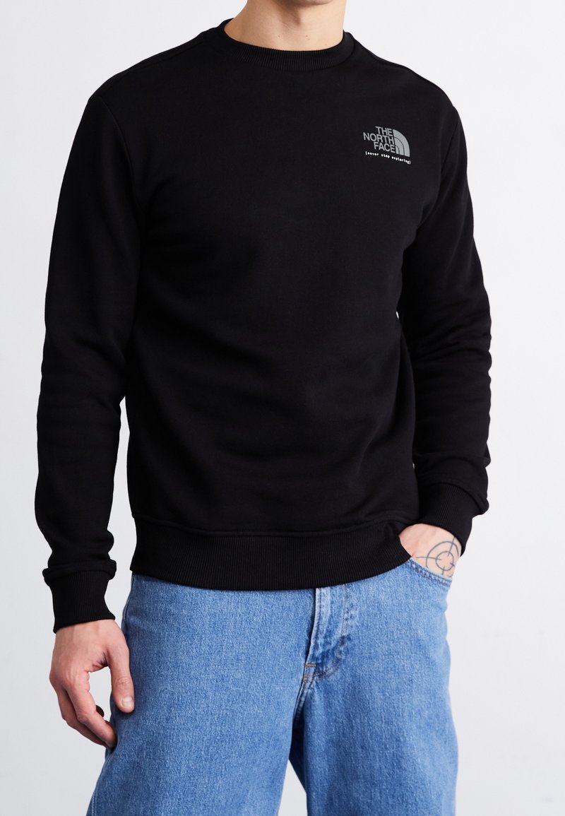 Personne portant un sweat-shirt noir The North Face et un jean bleu clair, avec un tatouage visible sur son poignet droit.