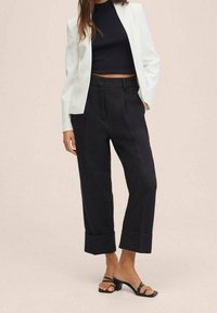 White tailored blazer over a black ribbed crop top, high-waisted black trousers with a wide leg and folded cuffs, and black strappy sandals.