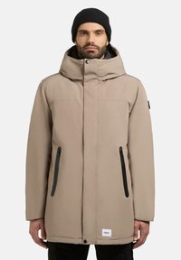 Beige parka-jakke med høj krave og hætte. Sorte lynlåslommer på siderne, blød tekstur og logodetalje ved kanten.