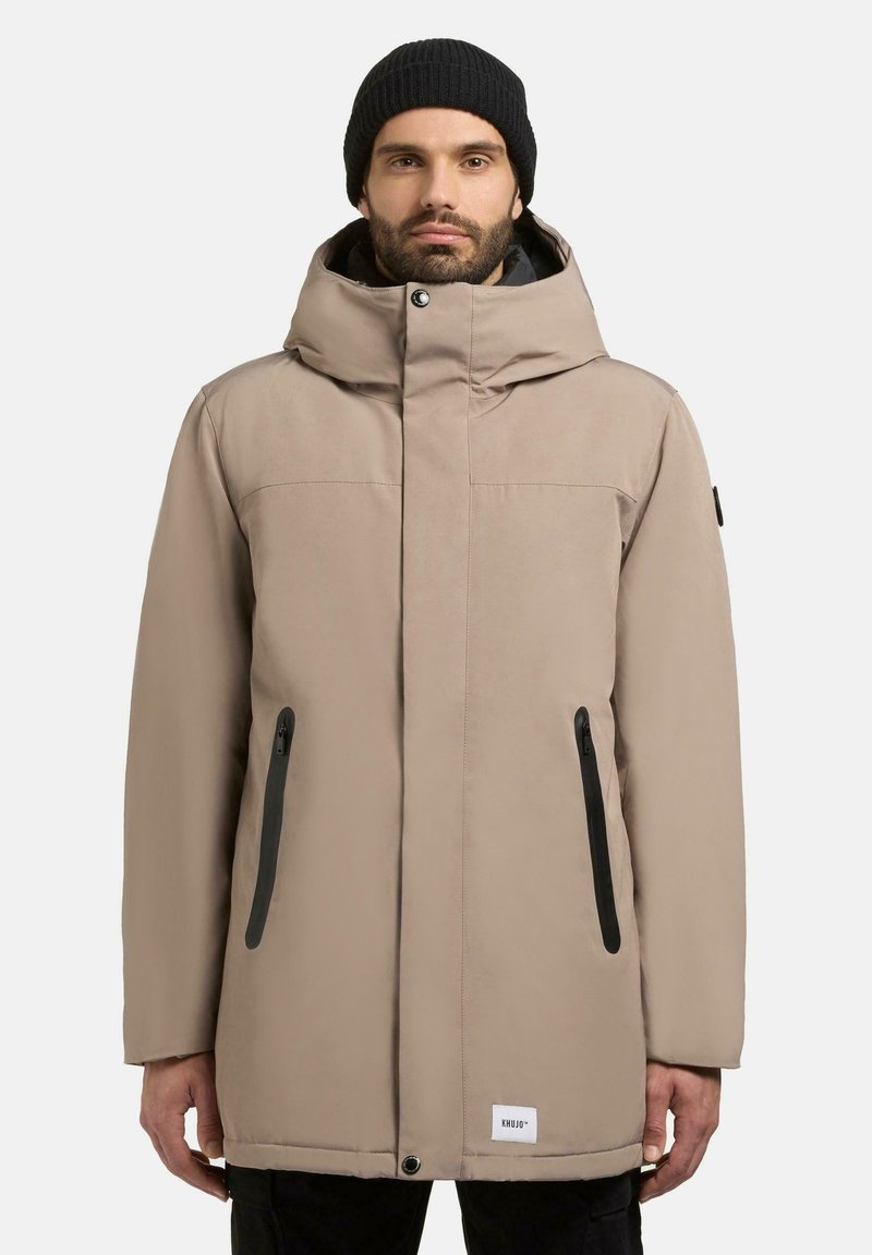 Beige parka-jakke med høj krave og hætte. Sorte lynlåslommer på siderne, blød tekstur og logodetalje ved kanten.