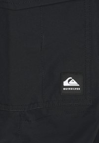 Svart tyg med en ficka som har en svart och vit Quiksilver-logopatch. Texturen är jämn med synliga sömdetaljer.